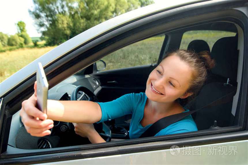 快乐的年轻女人拿着手机,开车时拍照. 微笑的女孩在车